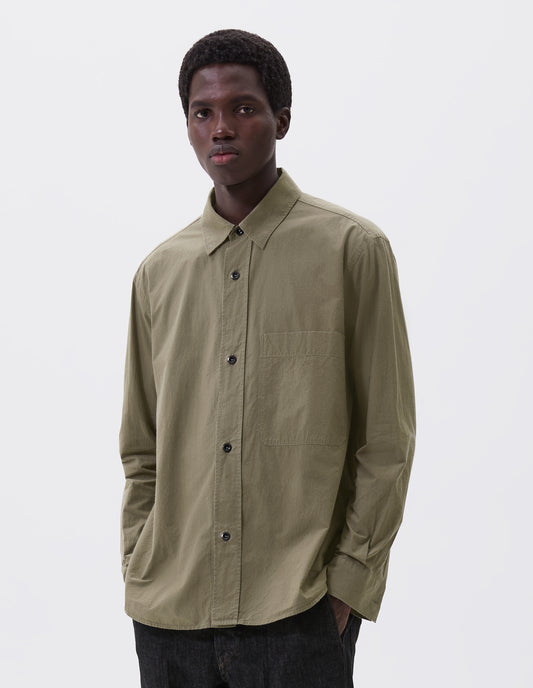 Cropped body image, front view of model wearing green brushed cotton shirt with raised placket, large chest pocket, corozo button closure and side gusset stitch detailing. Styled with black denim jeans.