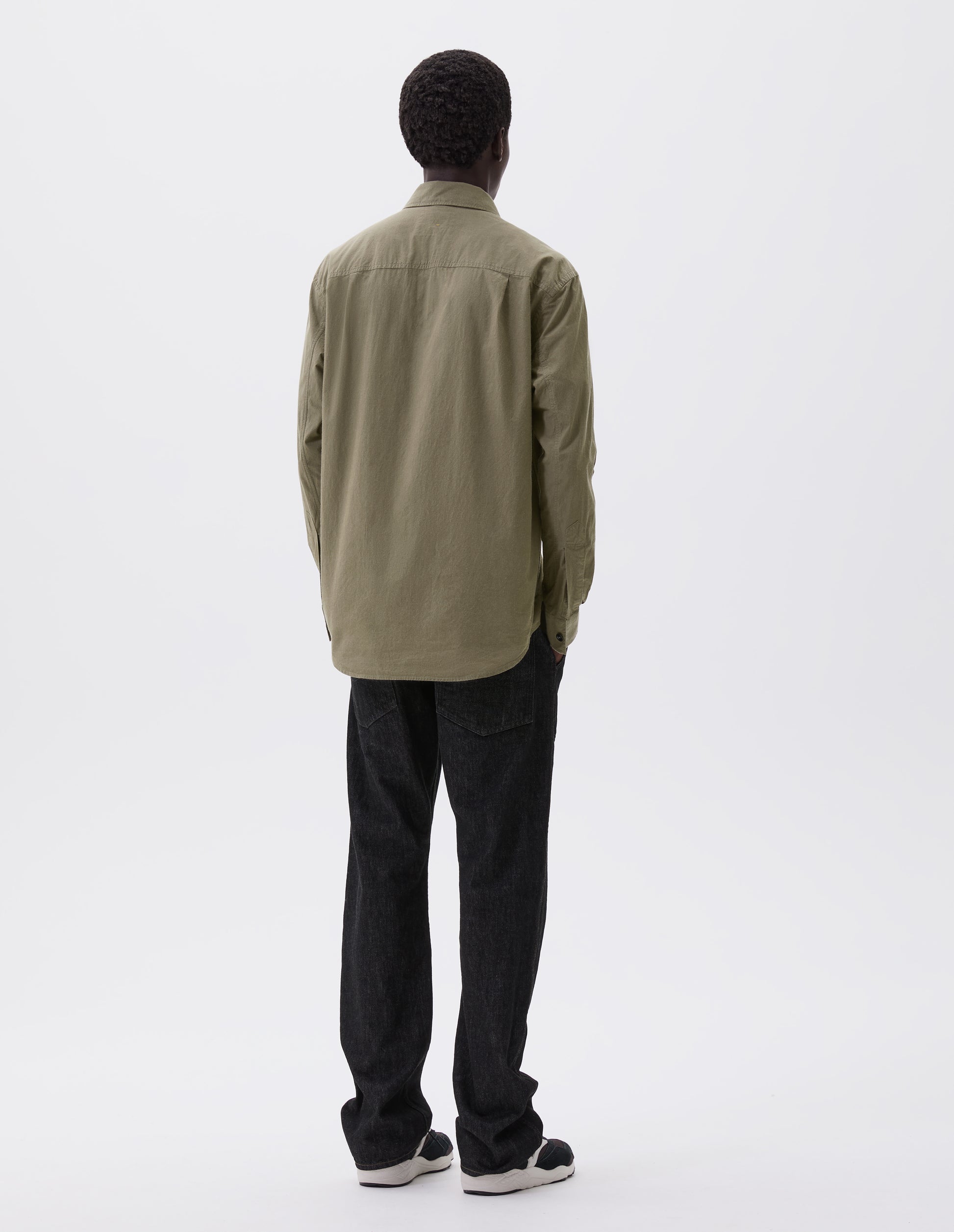 Full body image, back view of model wearing green brushed cotton shirt with side gusset stitch detailing and pleated back yoke. Styled with black denim jeans and black and brown trainers.