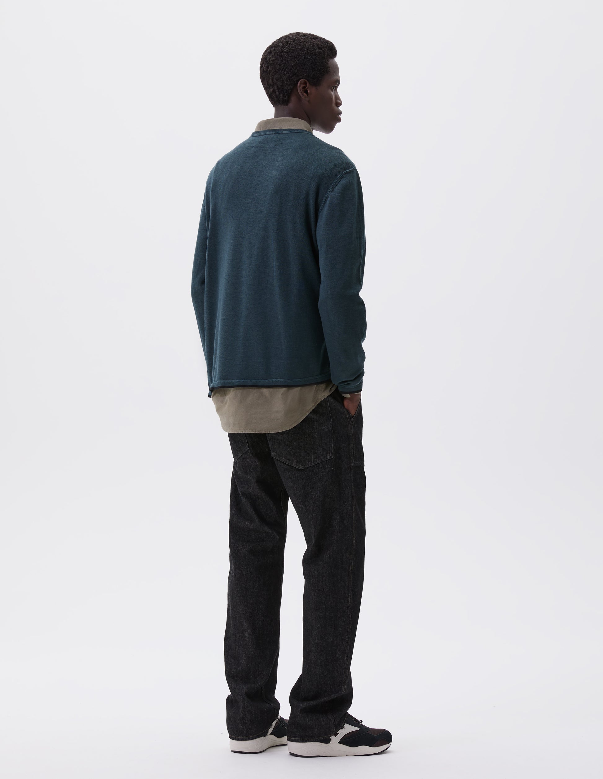 Full body image, front view of model wearing black denim twill jeans, with metal button fly, grown on waistband with belt loops and back pockets with half cotton lining. Styled with green shirt, blue jumper and black and brown trainers.