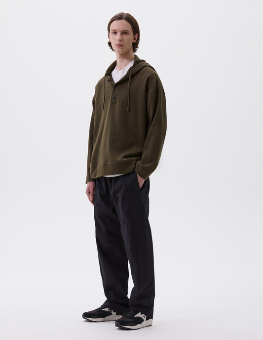 Full body image, front view of model wearing green cotton hoodie, with drawcord hood, corozo buttons, straight cuffs and stitch seam detailing. Styled with white t-shirt, indigo blue trousers and brown and black trainers.