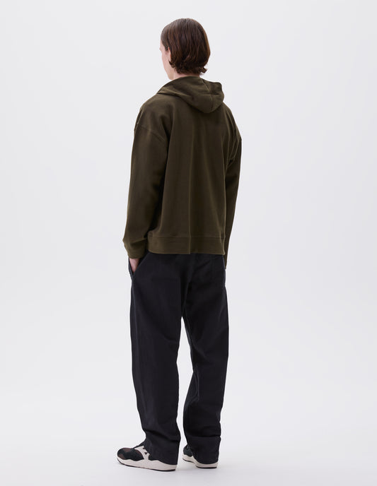 Full body image, back view of model wearing green cotton hoodie, with drawcord hood, corozo buttons, straight cuffs and stitch seam detailing. Styled wit indigo blue trousers and brown and black trainers.