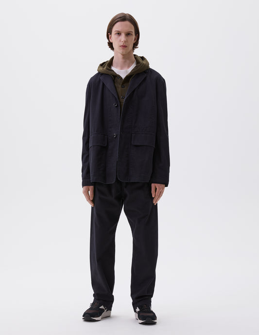 Full body image, front view of model wearing indigo blue twill blazer with corozo button fastening lapel, stitch through chest pocket and flap patch pockets. Styled over white t-shirt and green hooded sweatshirt, with matching indigo blue trousers and black and brown trainers.