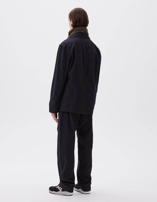 Full body image, back view of model wearing indigo blue twill blazer with back centre vent. Styled with green hooded sweatshirt, matching indigo blue trousers and black and brown trainers.
