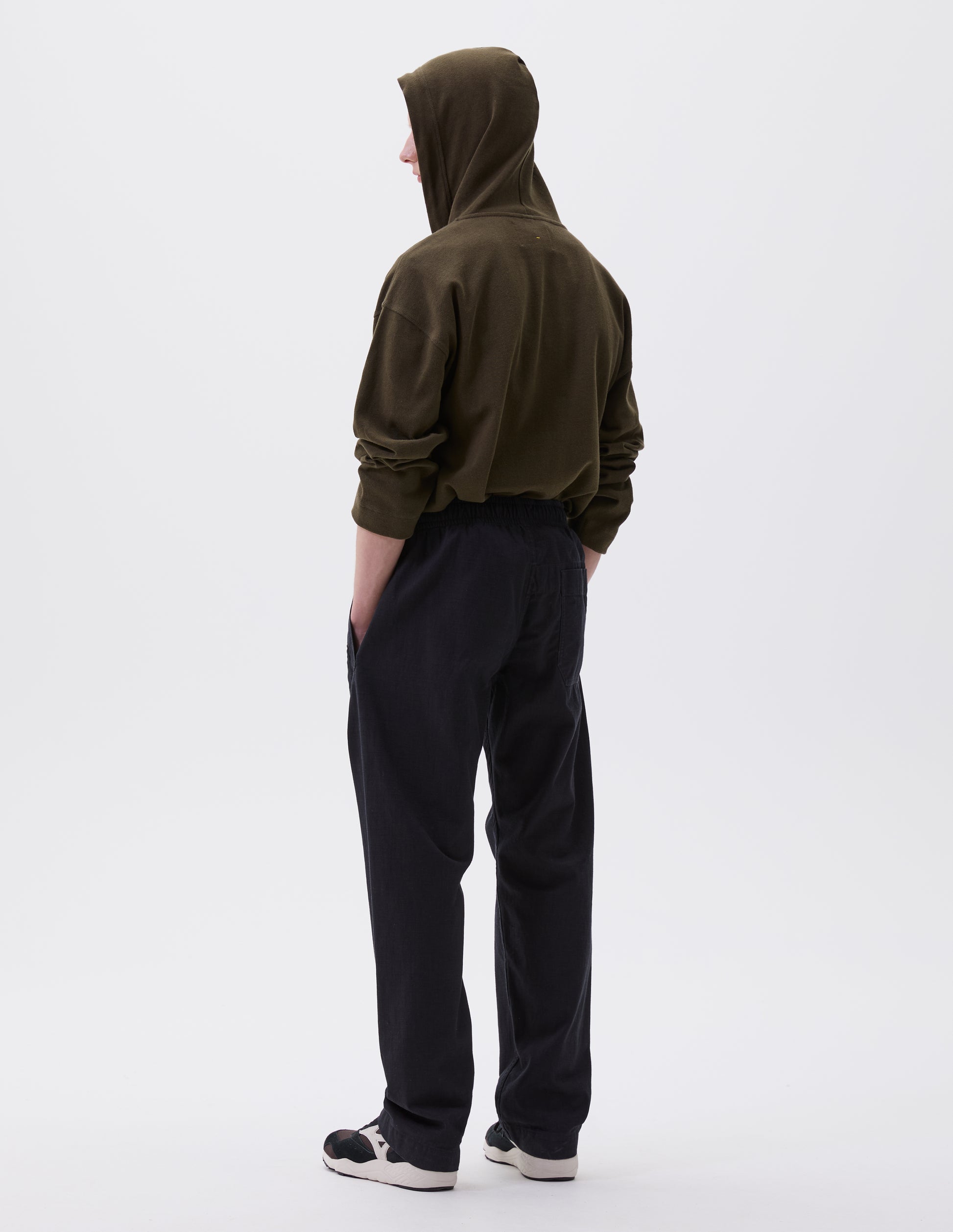 Full body image, back view of model wearing relaxed dark blue joggers with elasticated waist and patch back pocket. Styled with green hoodie and black, brown and white trainers.