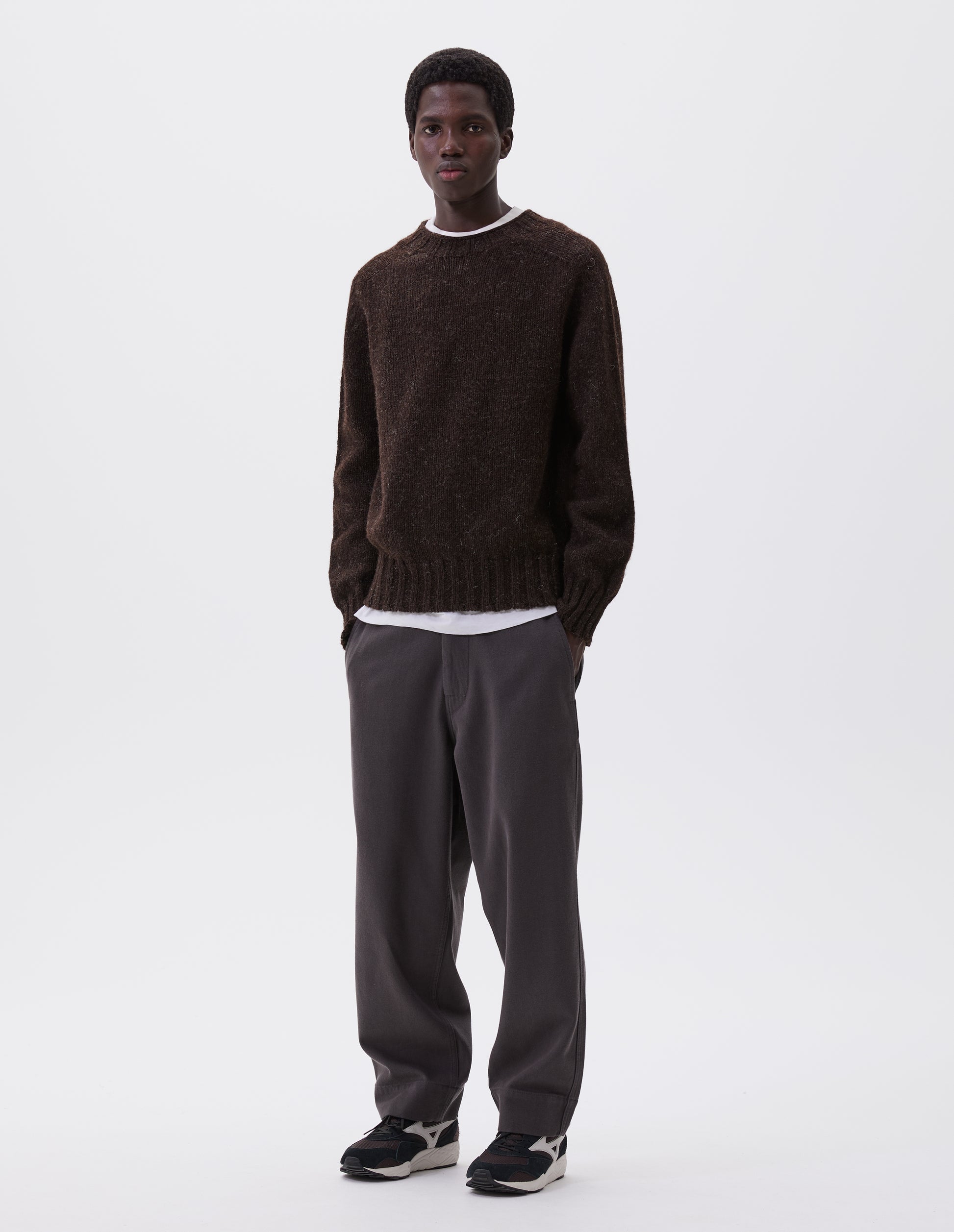 Full body image, front view of model wearing dark brown marl jumper with saddle sleeves and contrasting rib on neck, cuffs and hem. Styled over white t-shirt, with grey trousers and black and brown trainers.