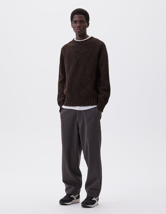 Full body image, front view of model wearing dark brown marl jumper with saddle sleeves and contrasting rib on neck, cuffs and hem. Styled over white t-shirt, with grey trousers and black and brown trainers.