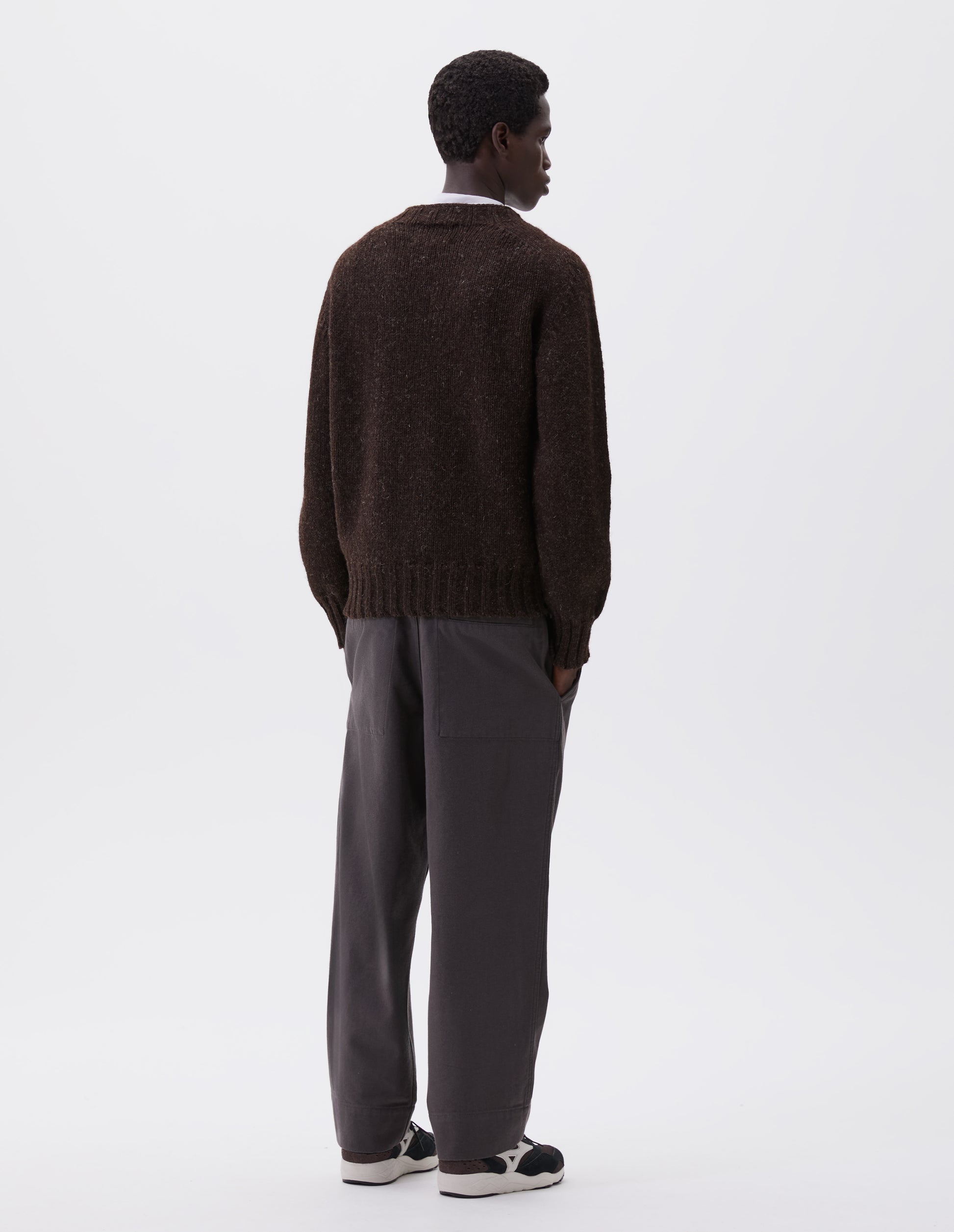 Full body image, back view of model wearing dark brown marl jumper with saddle sleeves and contrasting rib on neck, cuffs and hem. Styled over white t-shirt, with grey trousers and black and brown trainers.
