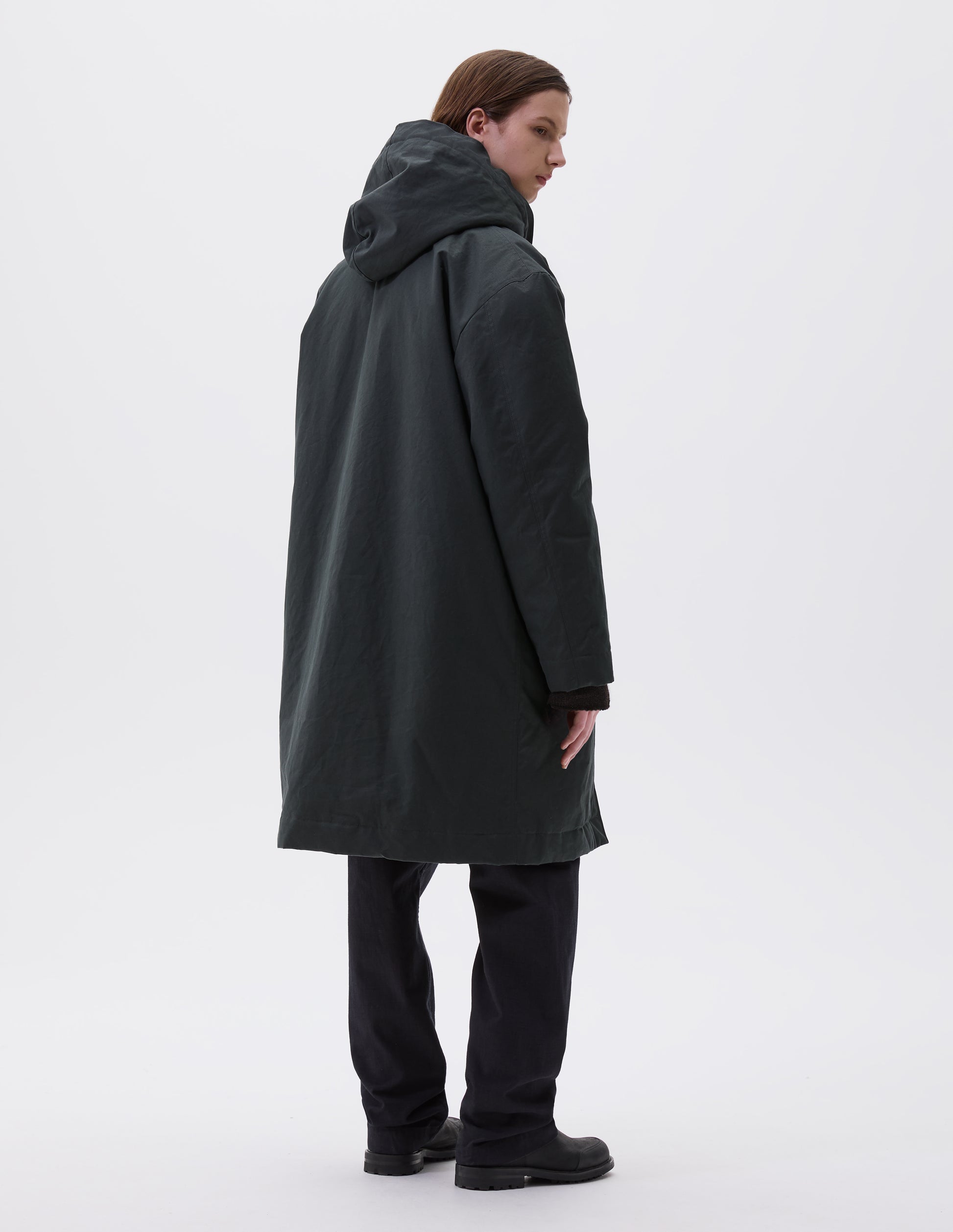 Full body image, back view of model wearing  blueish green hooded parka with fleece lined drawcord hood. Styled with indigo blue trousers and black leather boots.
