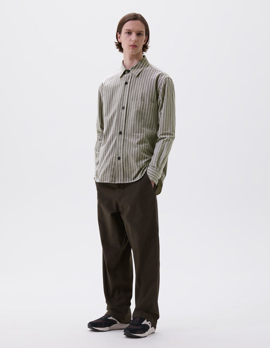 Full body image, front view of model wearing green, grey and white striped cotton long sleeve shirt, with raised placket, large chest pocket, corozo button closure and side gusset stitch detailing. Styled with green trousers and black and brown trainers.