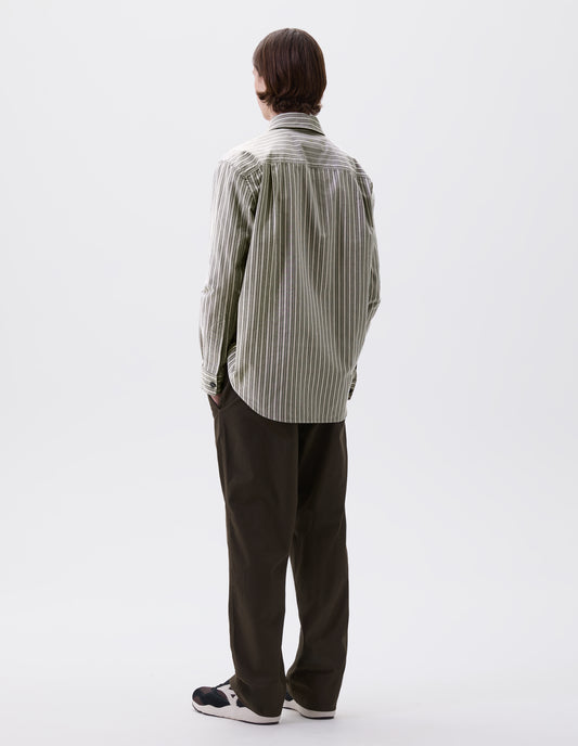 Full body image, back view of model wearing green, grey and white striped cotton long sleeve shirt, with side gusset stitch detailing and pleated back yoke. Styled with green trousers and black and brown trainers.