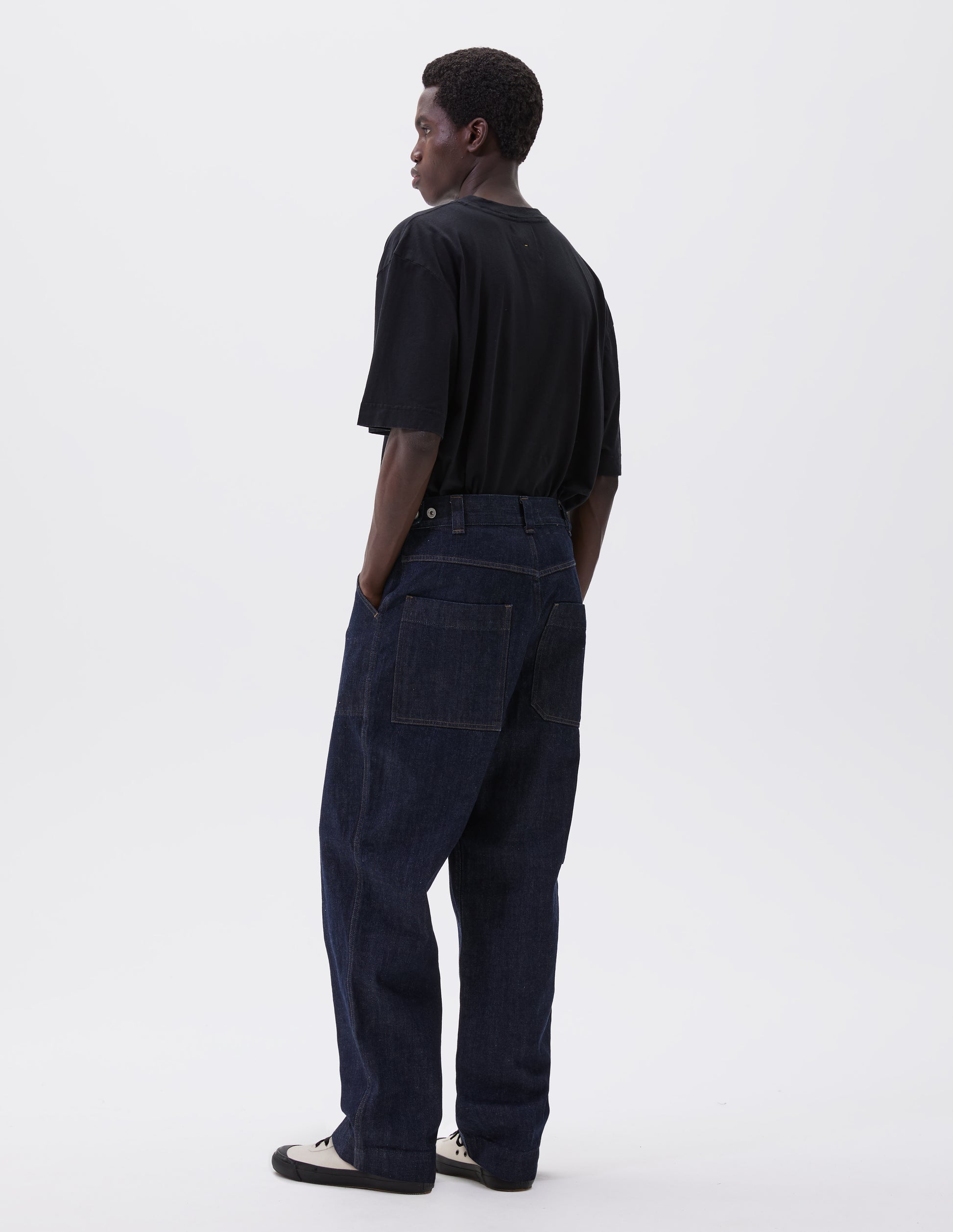 Full body image, back view of model wearing indigo blue denim twill jeans. With metal button tab waist adjusters, belt loops and patch and hammer pocket on back of trousers. Styled with navy blue t-shirt and white canvas trainers.