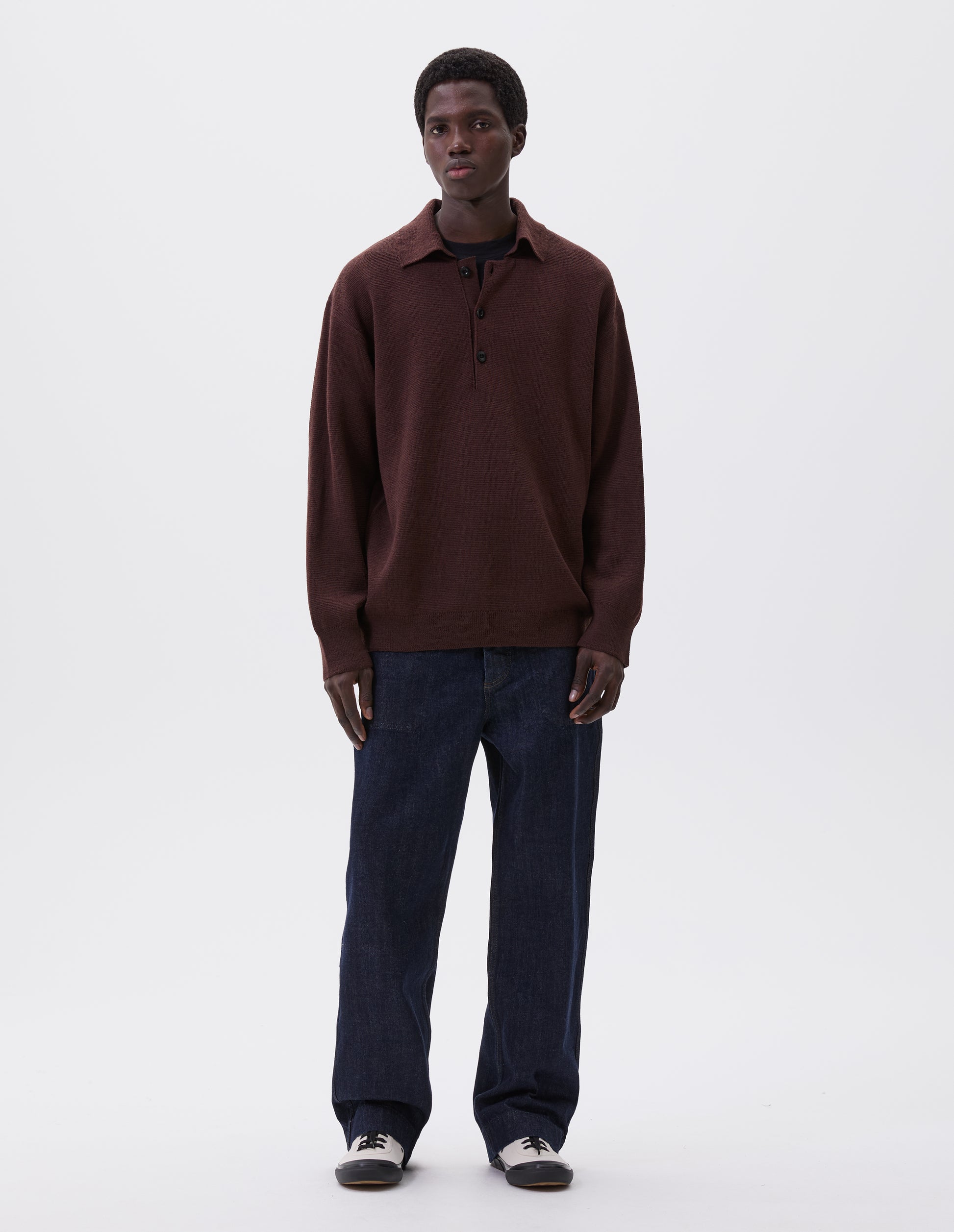 Full body image, front view of male model wearing a pair of white cotton canvas trainers with metal eyelet, black laces and contrast vulcanised rubber sole. Styled with burgundy knitted polo top and indigo blue jeans.