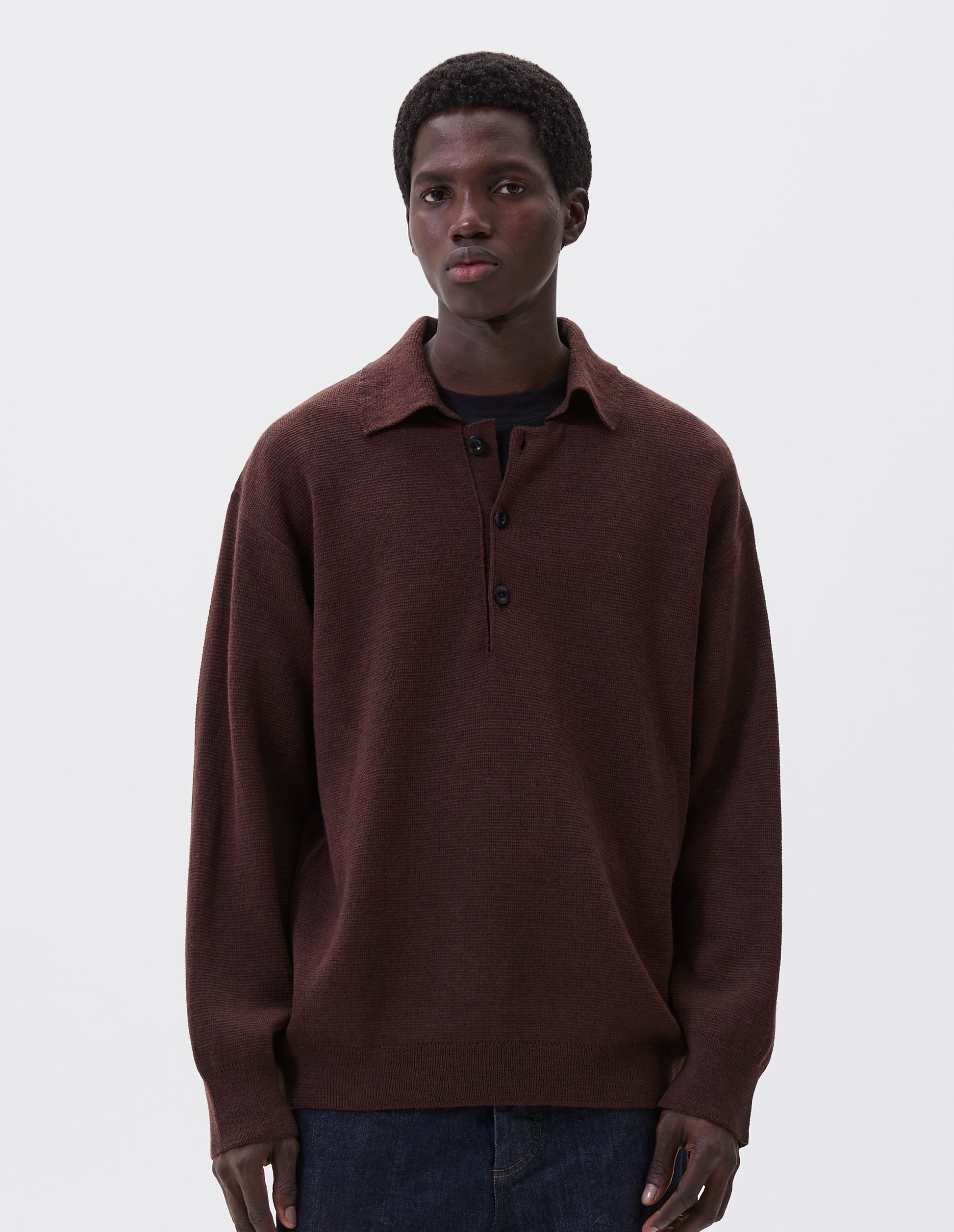 Cropped body image, front view of model wearing brown knitted polo with contrast rib on neck, cuffs and hem and corozo brace buttons. Styled over black t-shirt, with indigo blue jeans.