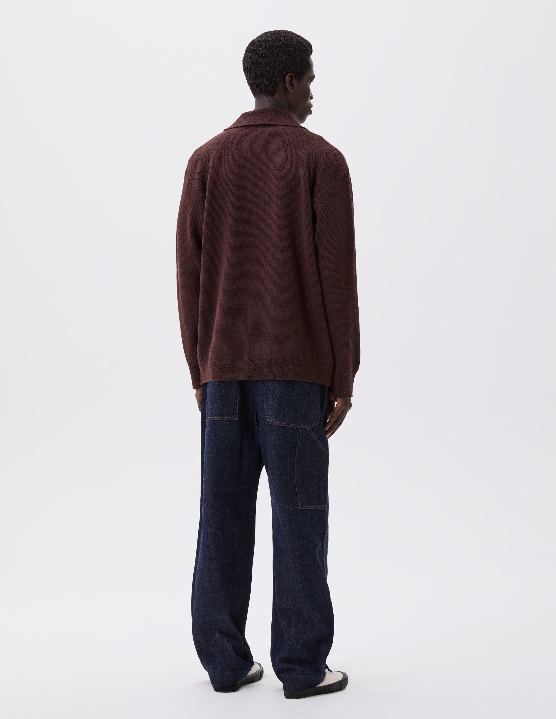 Full body image, back view of model wearing brown knitted polo with contrast rib on neck, cuffs and hem. Styled with indigo blue jeans and white canvas trainers. 