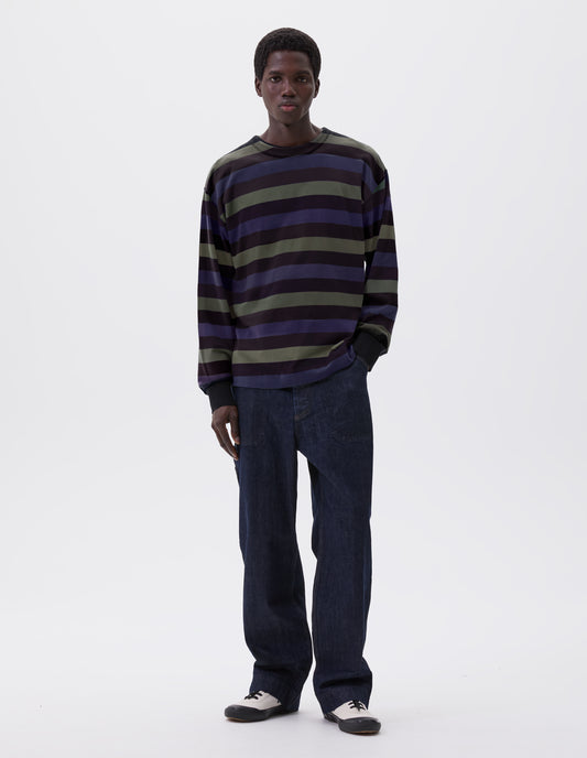 Full body image, front view of model wearing off black, blue and green striped long sleeve t-shirt, with single rib cuff and self faced neck. Styled with indigo blue jeans and ecru canvas trainers. 