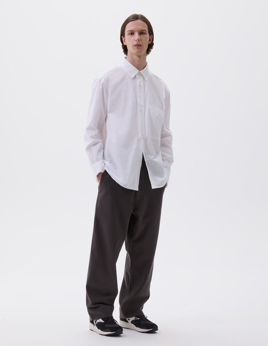 Full body image, front view of model wearing  relaxed white shirt with  raised placket, chest pocket and corozo brace button fastening. Styled with dark grey trousers and black, brown and white trainers.