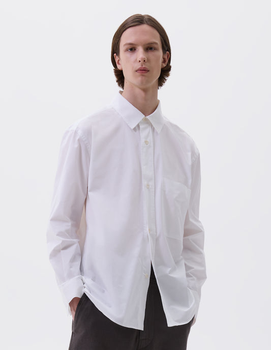 Cropped body image, front view of model wearing relaxed white shirt with  raised placket, chest pocket and corozo brace button fastening. Styled with dark grey trousers
