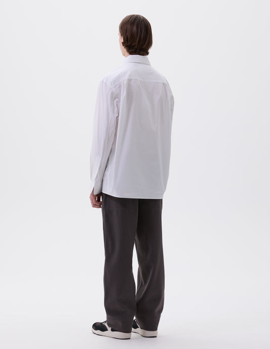 Full body image, back view of model wearing relaxed white shirt with pleats on back yoke and corozo brace buttons on cuffs. Styled with dark grey trousers and black, brown and white trainers.