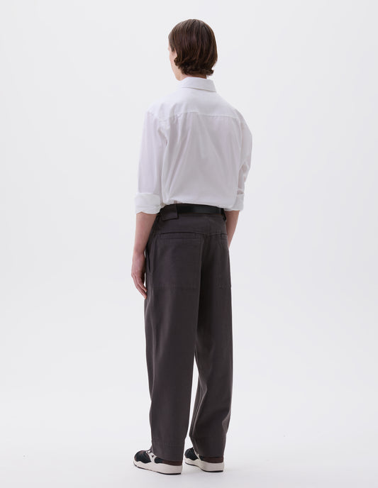Full body image, back view of model wearing grey trousers with side elasticated waistband, wide belt loops and stitch through welt back pockets on back yoke. Styled with white cotton shirt, black leather belt and black and brown trainers.