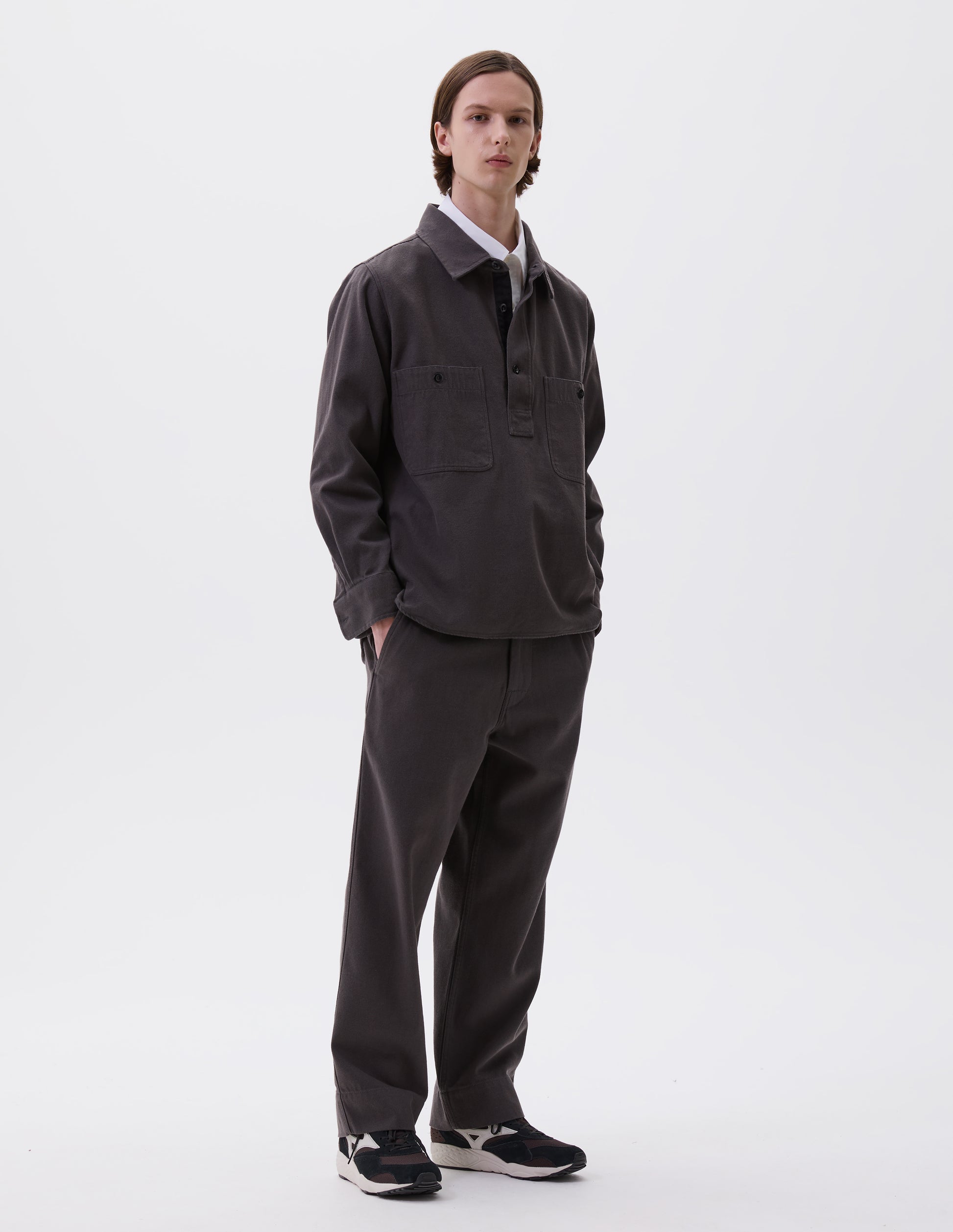 Full body image, front view of model wearing grey overhead shirt with raised placket, box stitch detailing, button through chest patch pockets and elongated back hem. Styled over white cotton shirt, with matching grey trousers and black and brown trainers.