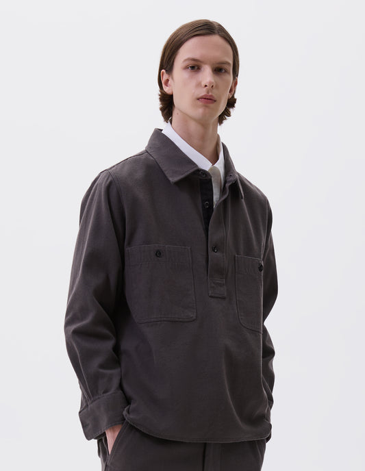 Cropped body image, front view of model wearing grey overhead shirt with raised placket, box stitch detailing, button through chest patch pockets and elongated back hem. Styled over white cotton shirt, with matching grey trousers.