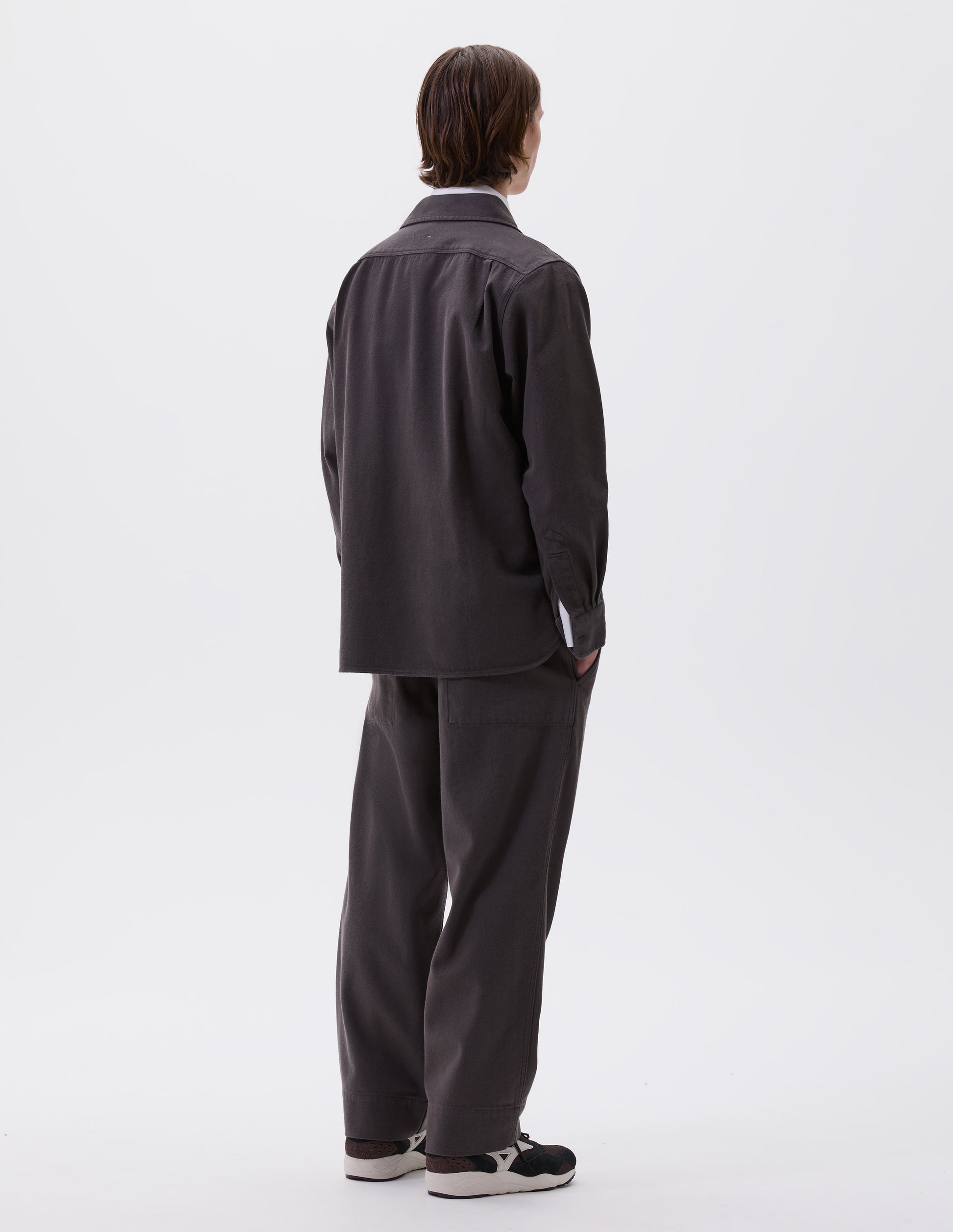 Full body image, back view of model wearing grey cotton wool drill overhead shirt. Styled over white cotton shirt, with matching grey trousers and black and brown trainers.