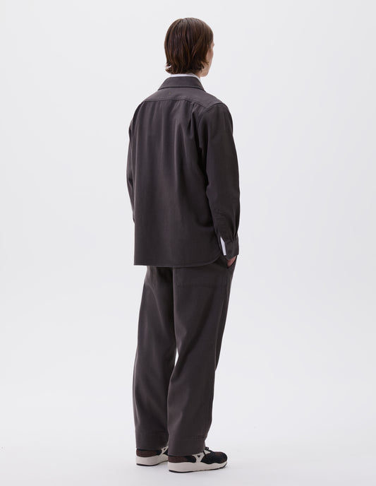 Full body image, back view of model wearing grey cotton wool drill overhead shirt. Styled over white cotton shirt, with matching grey trousers and black and brown trainers.