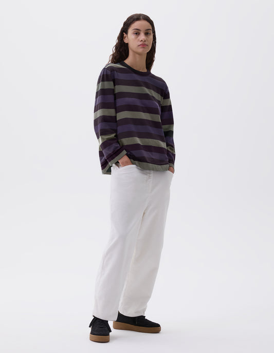 Full body image, front view of model wearing off black, blue and green striped long sleeve top, with contrast rib neck. Styled with white corduroy trousers and black nubuck leather trainers. 
