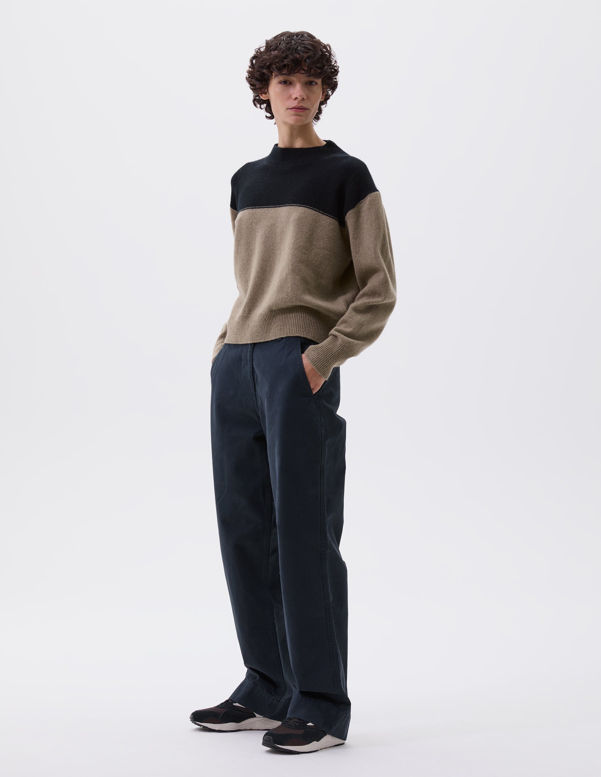 Full body image, front view of model wearing cropped black and beige jumper with ribbed crew neck, cuffs and hem. Styled with black trousers and black, brown and white trainers.