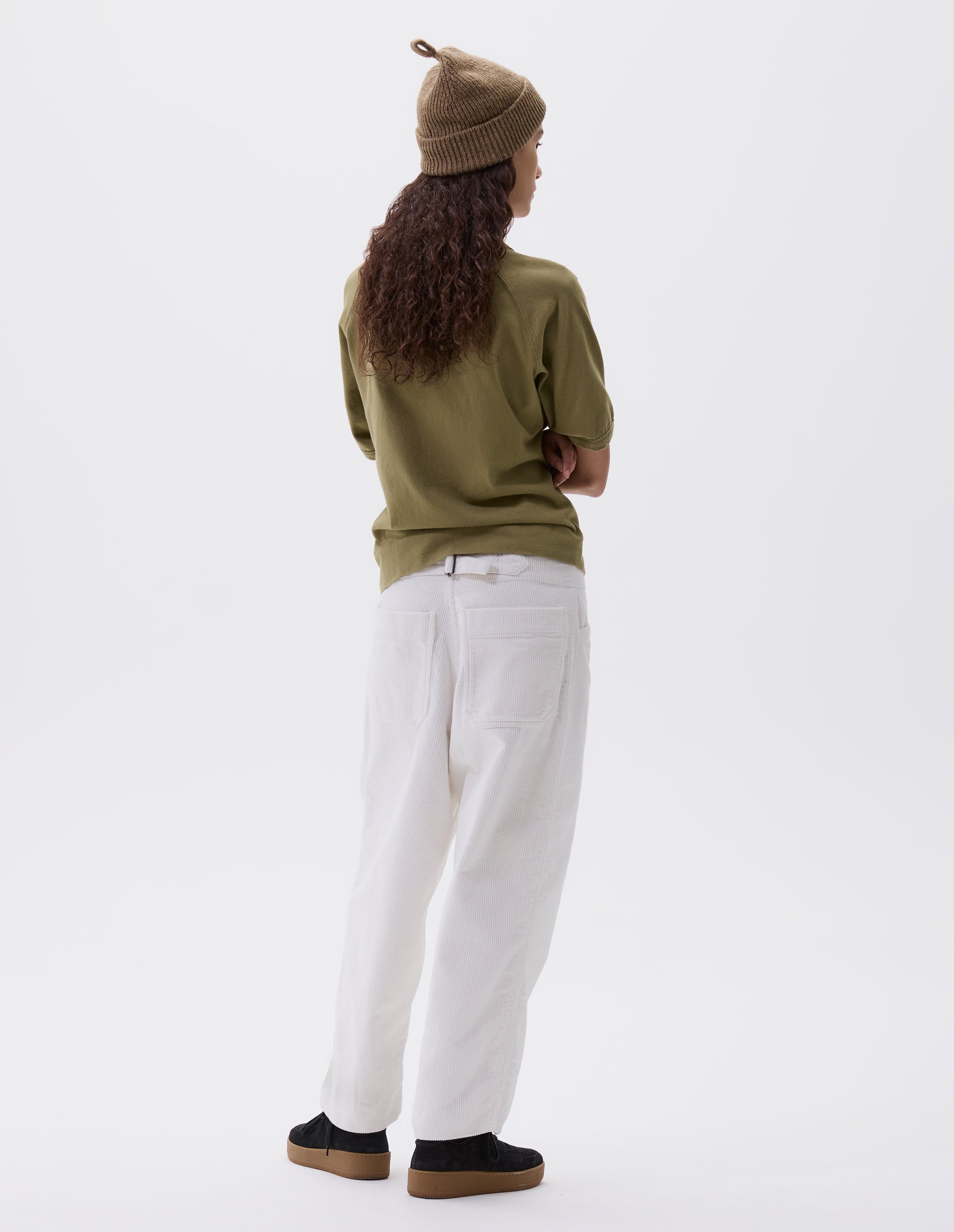 Full body image, back view of model wearing white heavy corduroy trousers with frog pockets, deep stitch-through hem, patch back pockets and cinch waist adjusters. Styled with beige knitted hat, green t-shirt and black suede trainers.