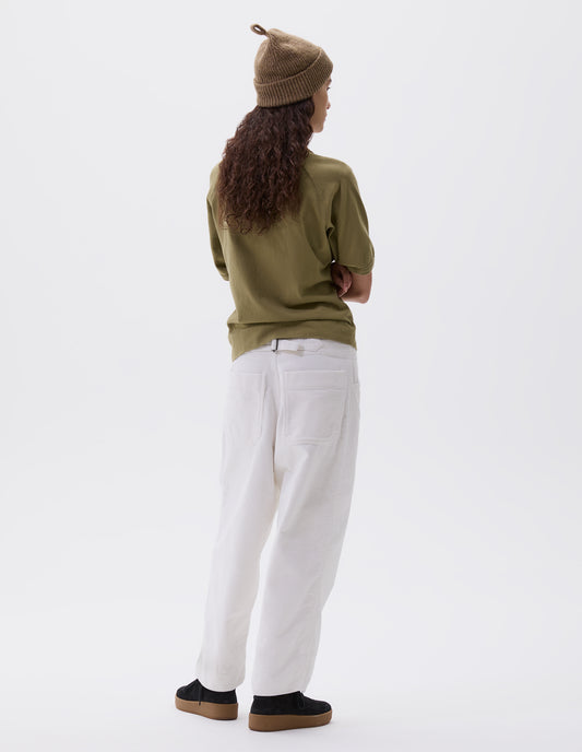 Full body image, back view of model wearing white heavy corduroy trousers with frog pockets, deep stitch-through hem, patch back pockets and cinch waist adjusters. Styled with beige knitted hat, green t-shirt and black suede trainers.