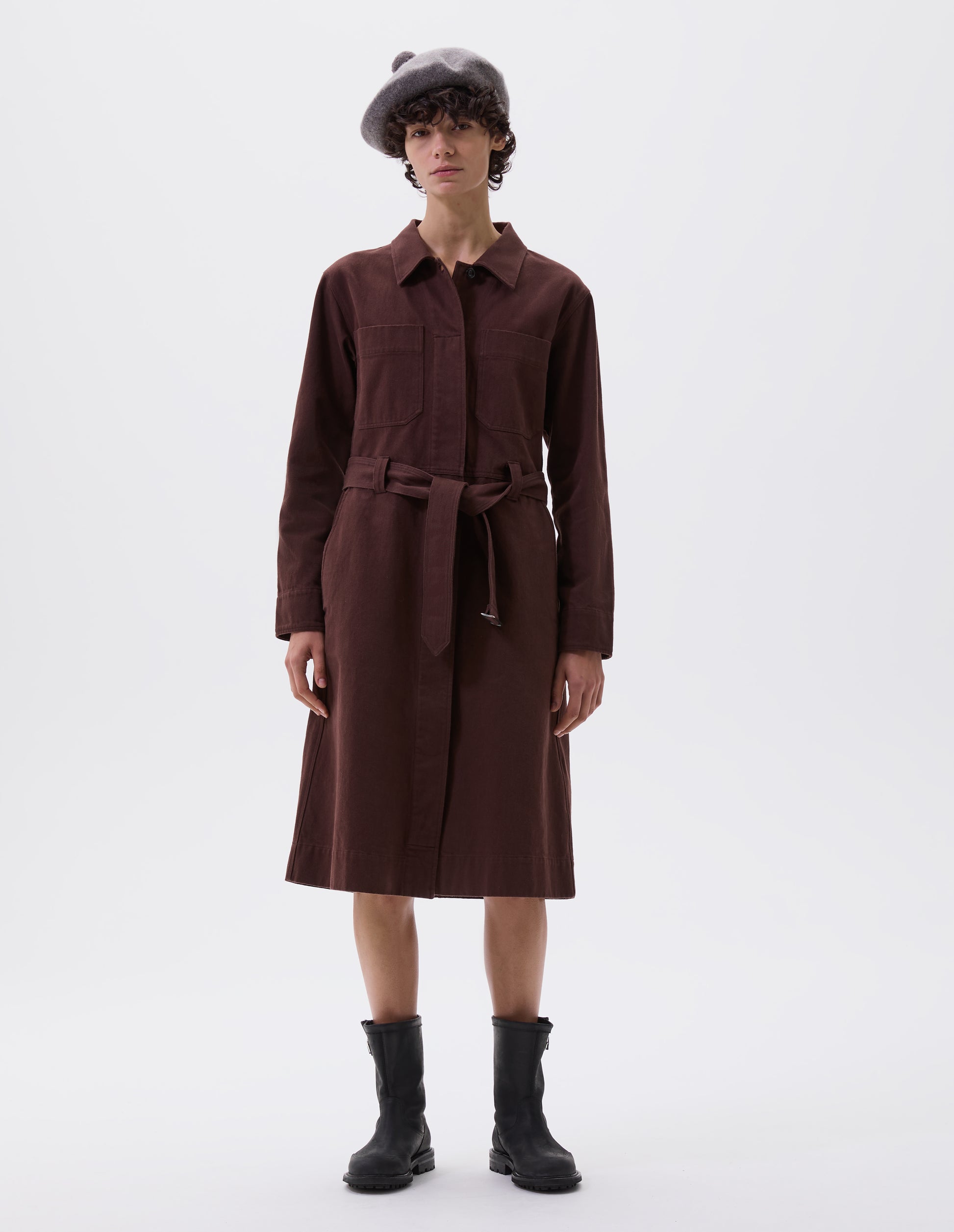 Full body image, front view of model wearing brown red long sleeve shirt dress, with fly front corozo button fastening, patch chest and slant side pockets, button fastening cuffs and removeable belt with metal slide adjuster. Styled with grey bobble hat and black leather boots.