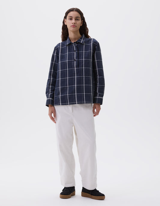 Full body image, front view of model wearing navy and white checked shirt with raised placket, corozo brace buttons, large dropped check patch pocket and gusset stitching. Styled with white corduroy trousers and black suede trainers.