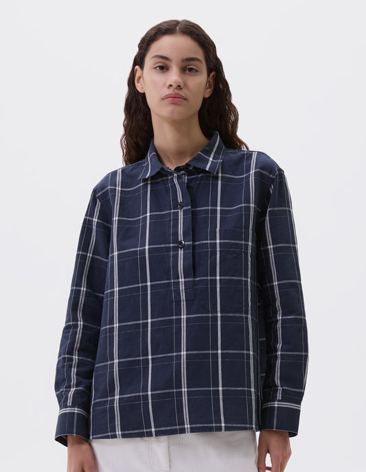 Cropped body image, front view of model wearing navy and white checked shirt with raised placket, corozo brace buttons, large dropped check patch pocket and gusset stitching. Styled with white corduroy trousers.