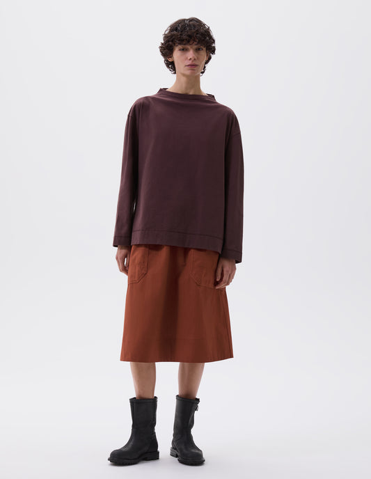 Full body image, front view of model wearing dark brown smock neck long sleeve t-shirt. Styled with dark orange skirt with large patch pockets and black leather biker boots.