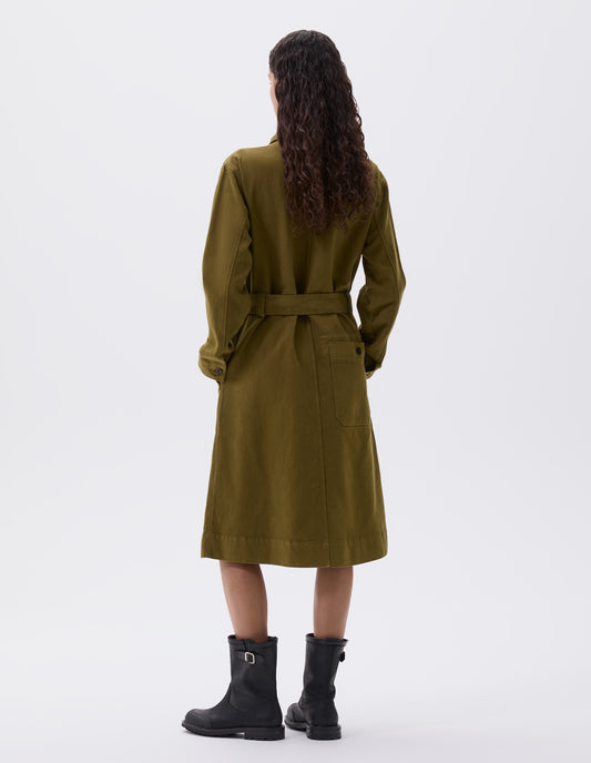 Full body image, back view of model wearing olive green long sleeve shirt dress, patch back pocket, corozo button fastening cuffs and removeable belt. Styled with black leather boots. 