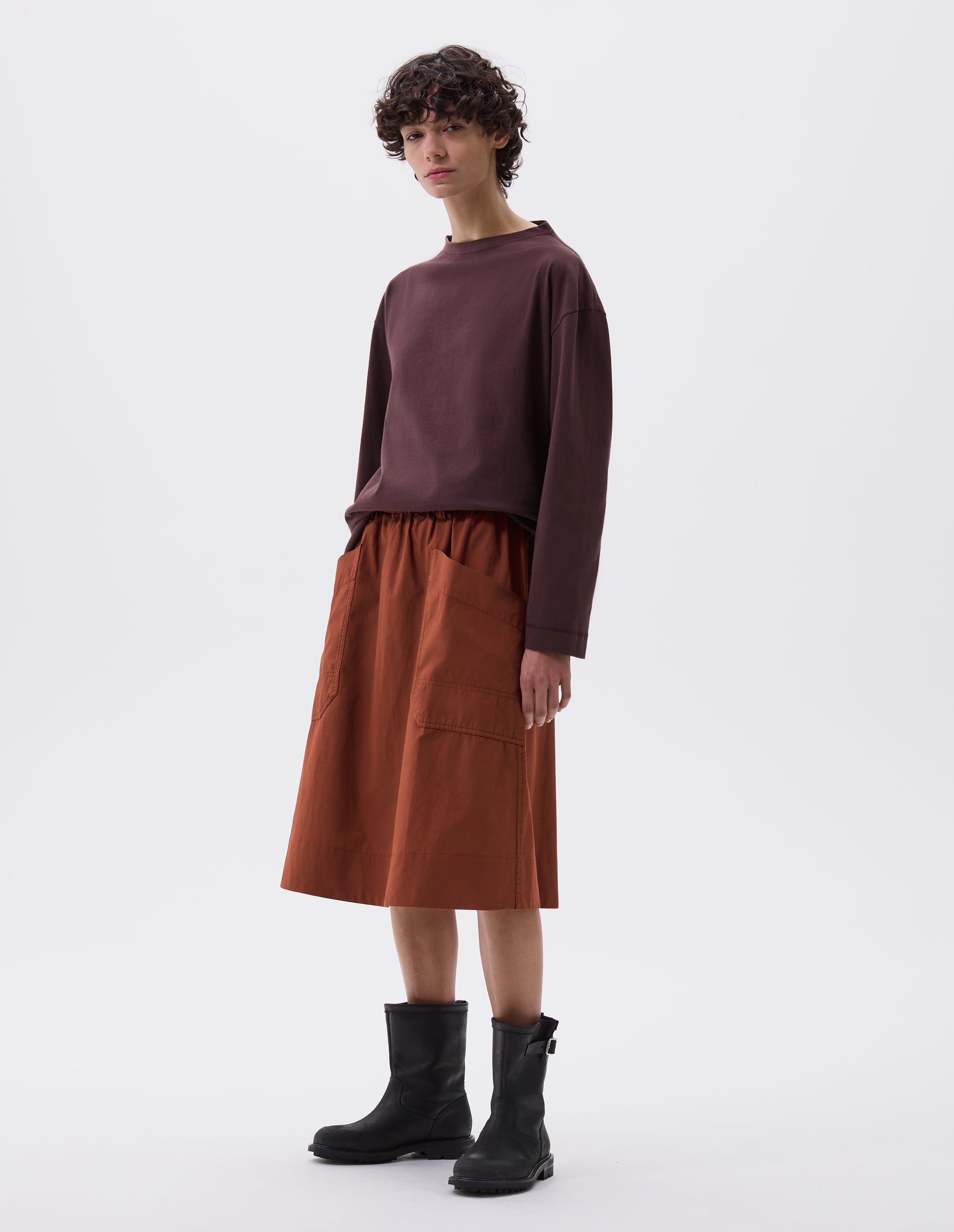 Full body image, front view of model wearing dark orange relaxed midi A-line skirt with elasticated waist and large, slant patch pockets. Styled with dark purple long sleeve t-shirt and black leather biker boots.