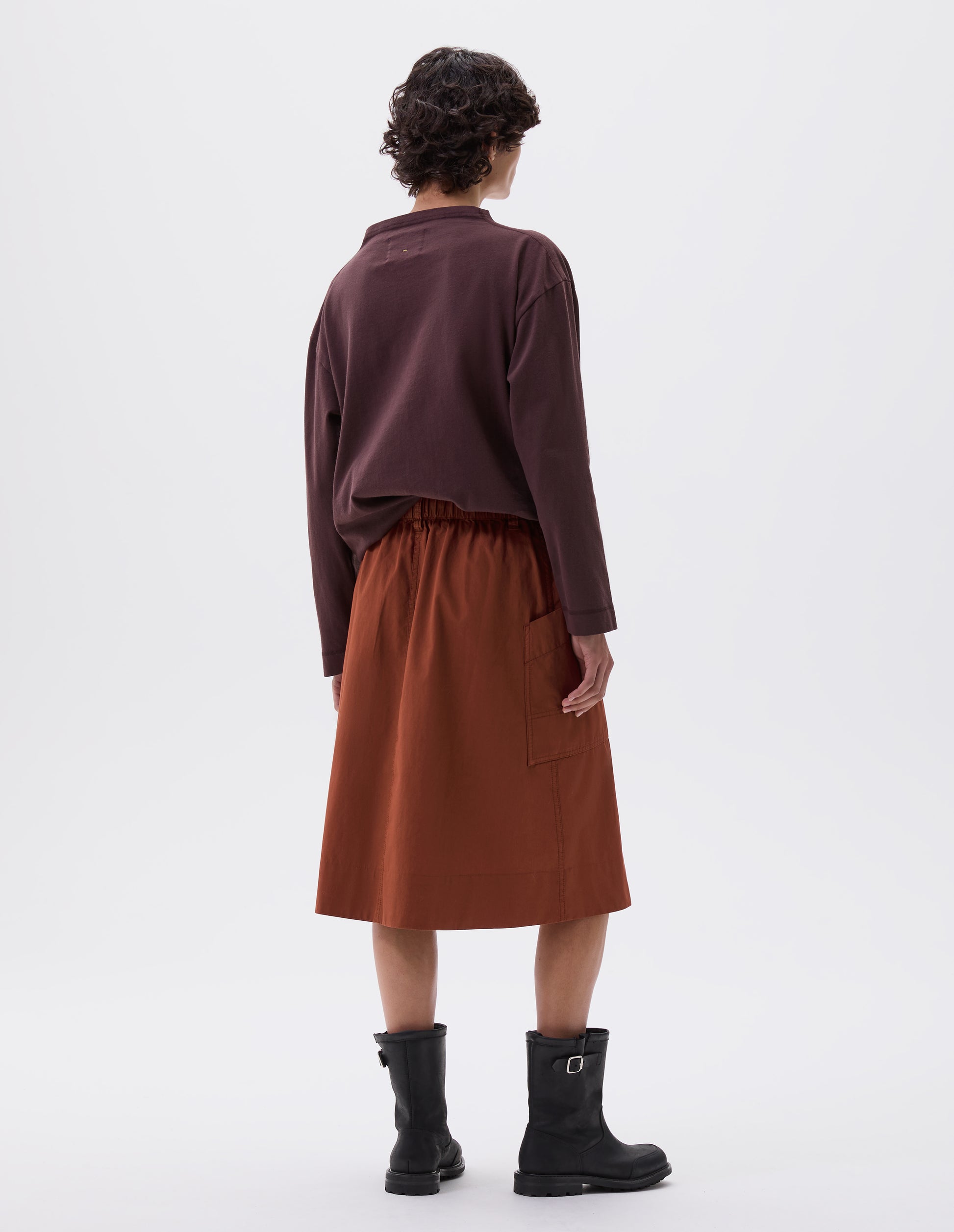 Full body image, back view of model wearing dark orange relaxed midi A-line skirt with elasticated waist and large, slant patch pockets. Styled with dark purple long sleeve t-shirt and black leather biker boots.