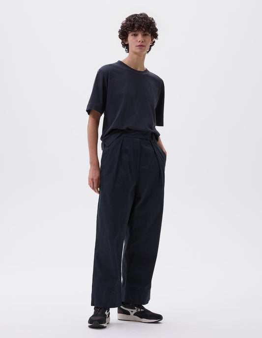 Full body image, front view of model wearing navy blue trousers with single pleat, concealed double hook and bar waist tab fastening, concealed side seam pockets and deep stitch hem. Styled with navy blue t-shirt and brown and black trainers. 