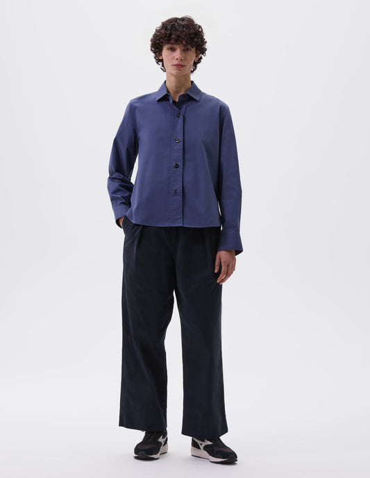 Full body image, front view of model wearing indigo blue shirt with raised placket and corozo button closure. Styled with navy blue trousers and brown and black trainers.