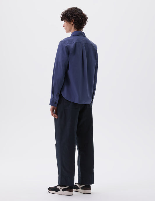 Full body image, back view of model wearing indigo blue shirt with box pleat on back yoke. Styled with navy blue trousers and brown and black trainers. 