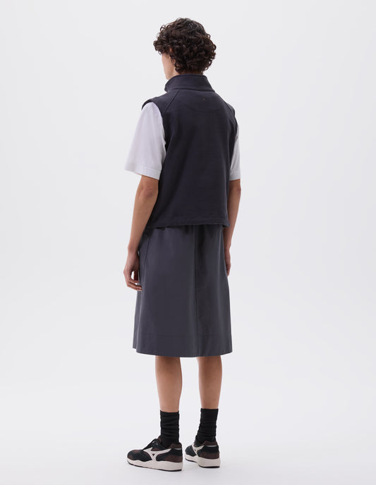 Full body image, back view of model wearing dark blue high neck sleeveless track top. Styled over white t-shirt and grey blue skirt, black socks and black, brown and white trainers. 