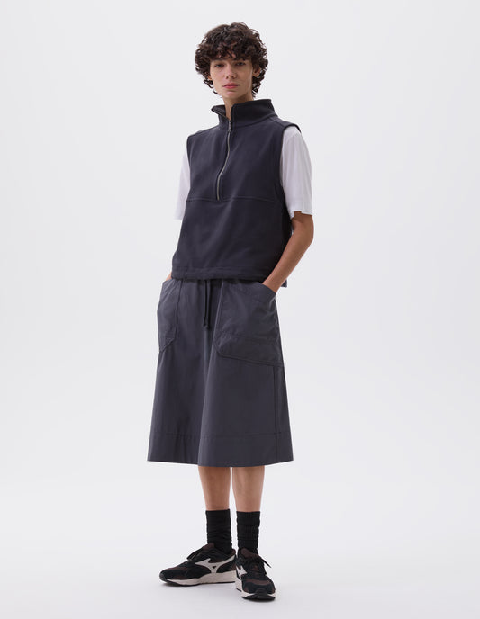 Full body image, front view of model wearing greyish blue relaxed midi A-line skirt with elasticated waist and large, slant patch pockets. Styled with dark blue sleeveless track top over white t-shirt, black socks with black, brown and white trainers.