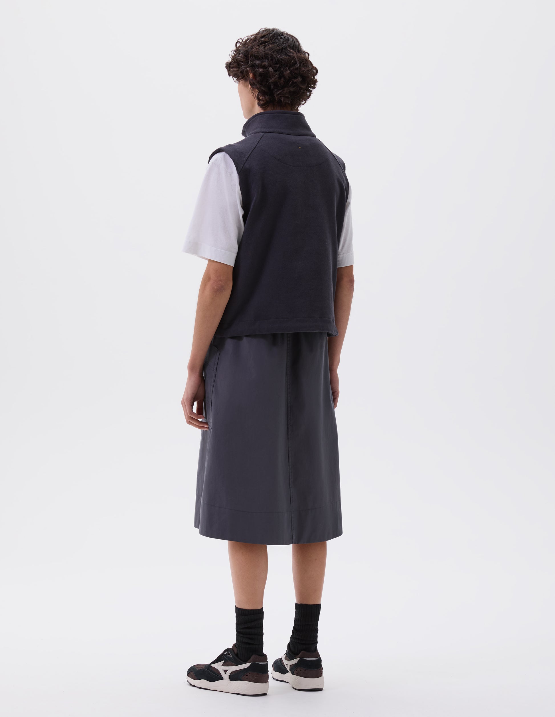 Full body image, back view of model wearing greyish blue relaxed midi A-line skirt with elasticated waist and large, slant patch pockets. Styled with dark blue sleeveless track top over white t-shirt, black socks with black, brown and white trainers.