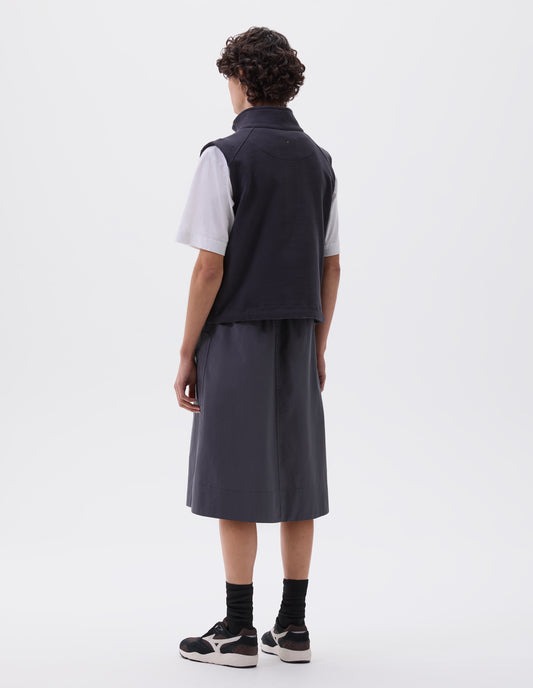 Full body image, back view of model wearing greyish blue relaxed midi A-line skirt with elasticated waist and large, slant patch pockets. Styled with dark blue sleeveless track top over white t-shirt, black socks with black, brown and white trainers.