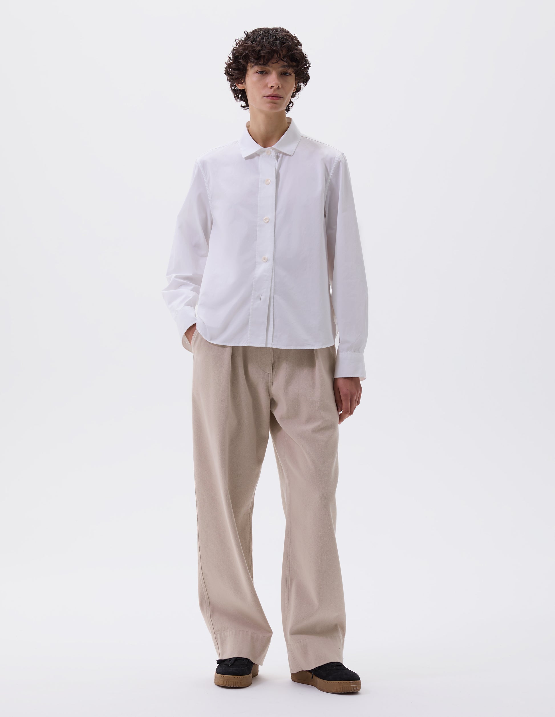 Full body image, front view of model wearing white regular cut shirt with stitch through raised placket, corozo buttons on placket and cuffs, curved hem. Styled with beige trousers and black nubuck trainers.