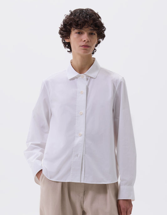 Cropped body image, front view of model wearing white regular cut shirt with stitch through raised placket, corozo buttons on placket and cuffs, curved hem. Styled with beige trousers. 