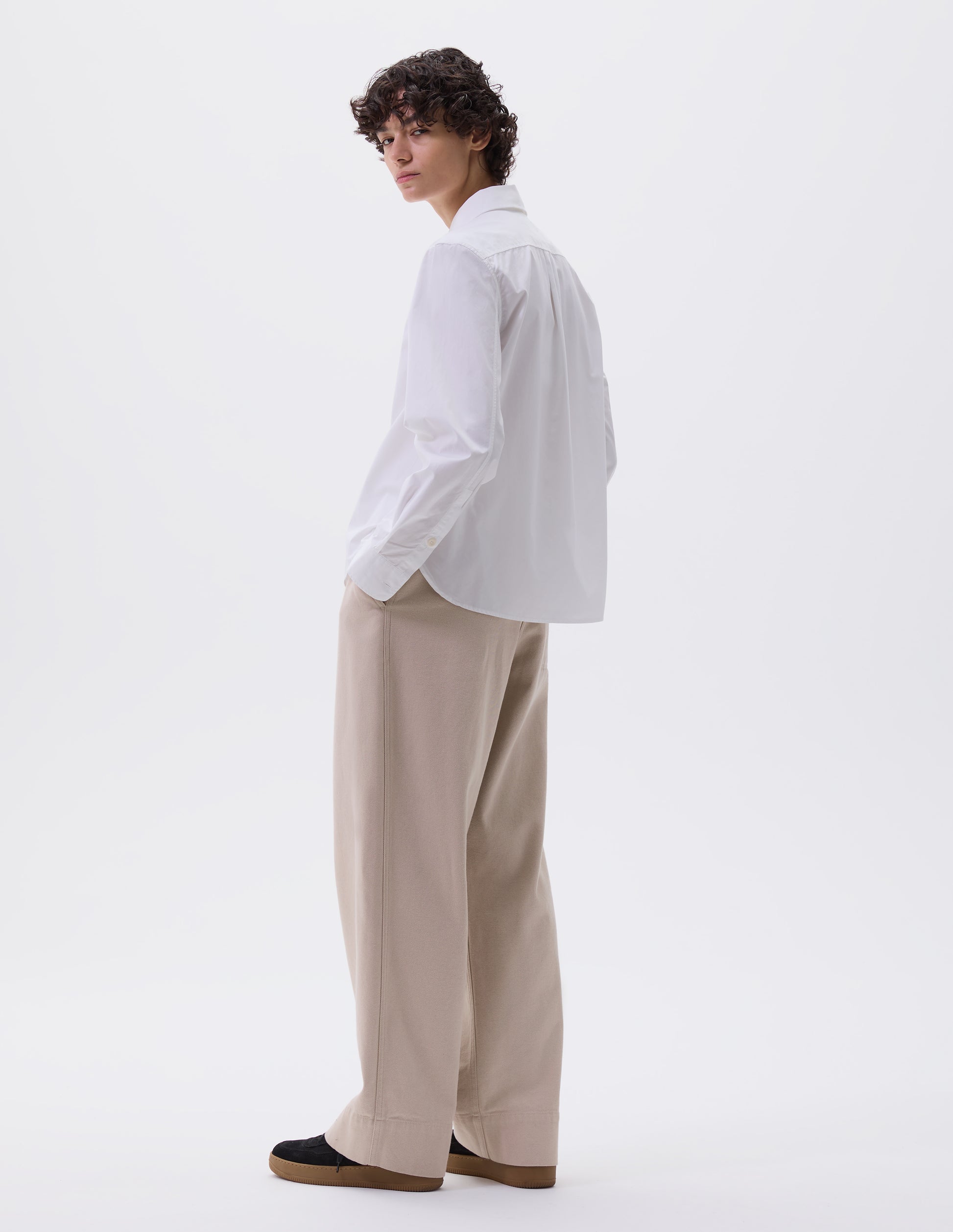 Full body image, back view of model wearing white regular cut shirt with box pleat on back yoke, corozo button cuffs and curved hem. Styled with beige trousers and black nubuck trainers.