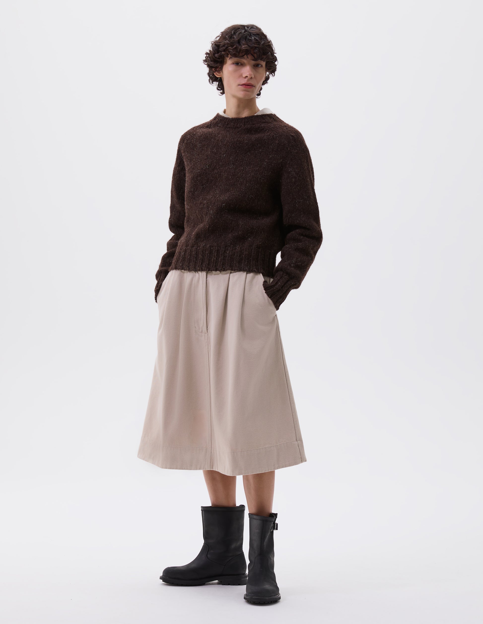 Full body image, front view of model wearing dark brown rustic wool jumper with saddle sleeve and contrasting rib on neck, cuffs and hem. Styled with beige skirt and black leather boots.