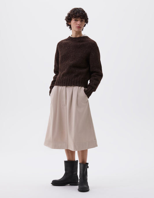 Full body image, front view of model wearing dark brown rustic wool jumper with saddle sleeve and contrasting rib on neck, cuffs and hem. Styled with beige skirt and black leather boots.